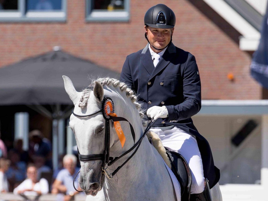 A man is smiling as he rides a grey horse in an arena, with spectators in the background.