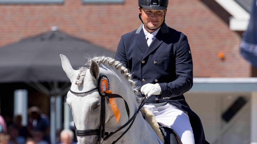 A man is smiling as he rides a grey horse in an arena, with spectators in the background.