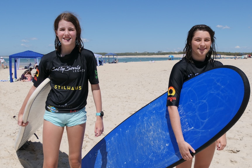 Two teenage girls in black rash vests stand smiling on a beach with surfboards under their arms.