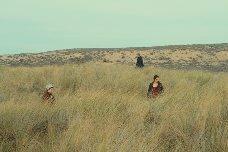 Three women in 18th century dresses wander through a field of tall grass.