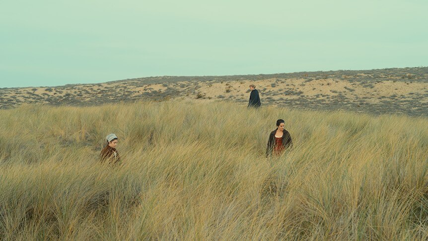 Three women in 18th century dresses wander through a field of tall grass.