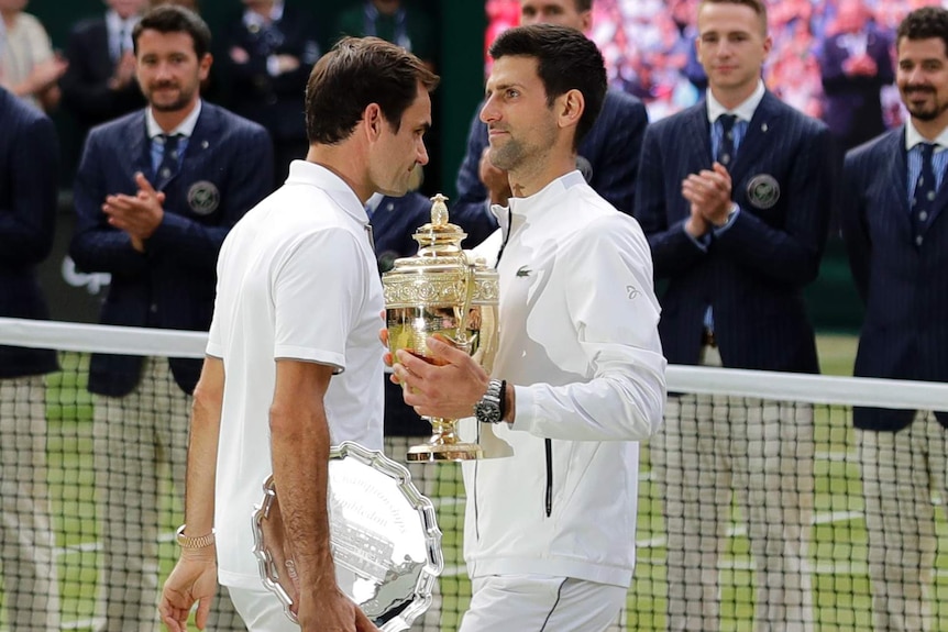 Novak Djokovic holds the Wimbledon trophy as Roger Federer walks past with the runner-up plate