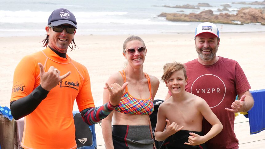 A male surf coach stands in a wetsuit, smiling, with a man and woman and their son, with the beach behind.