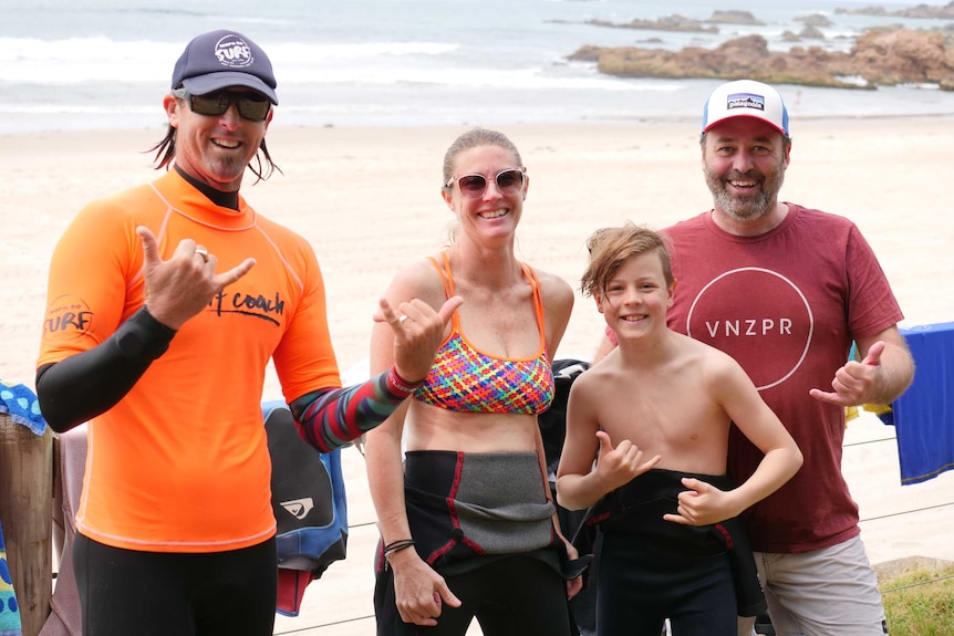 A male surf coach stands in a wetsuit, smiling, with a man and woman and their son, with the beach behind.