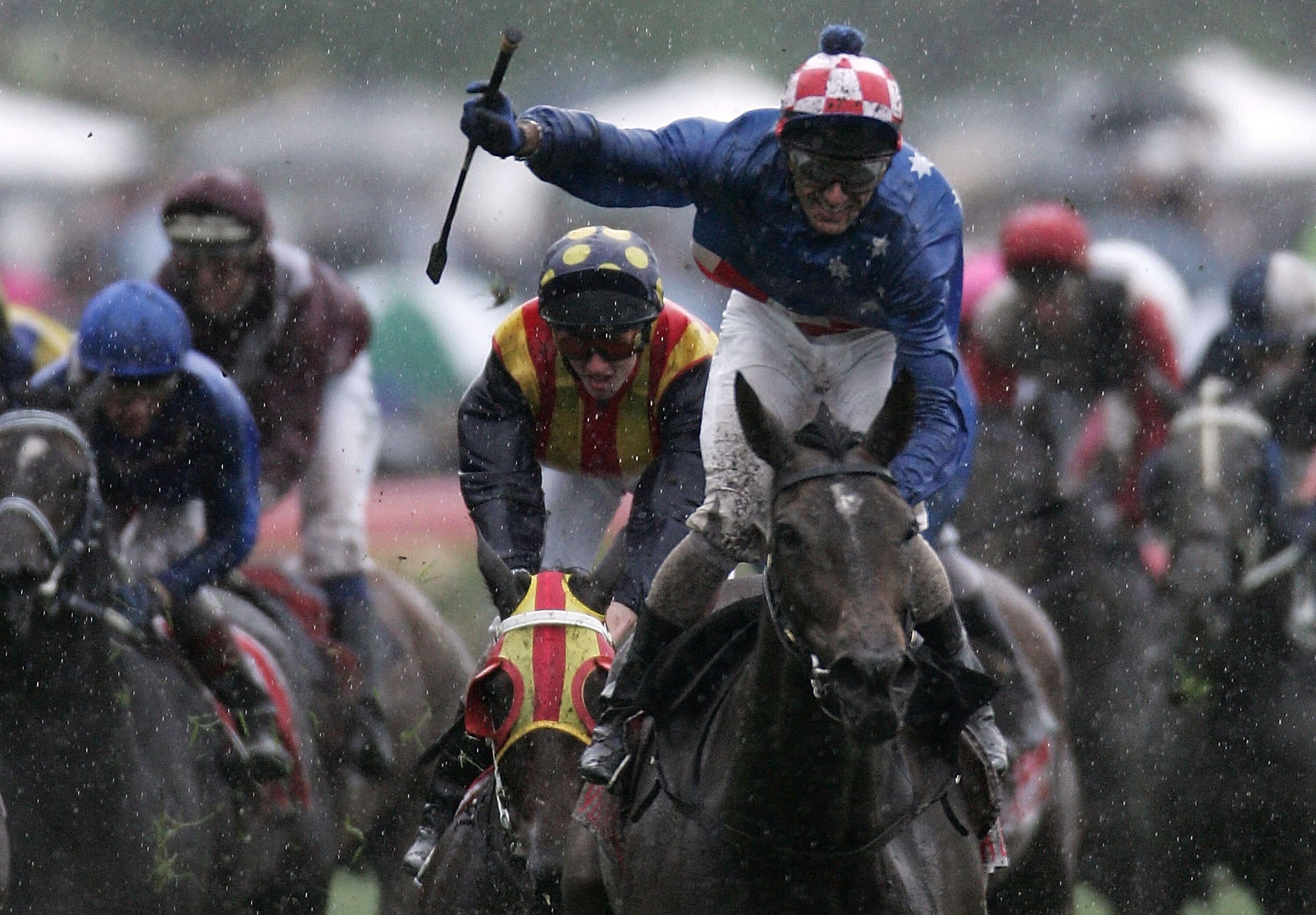 A head-on shot of a jockey standing up on his horse and brandishing his whip in celebration as the rain falls after a race.