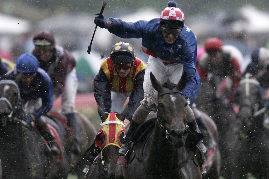 A head-on shot of a jockey standing up on his horse and brandishing his whip in celebration as the rain falls after a race.