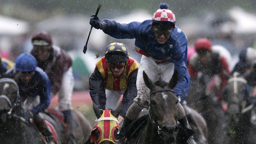 A head-on shot of a jockey standing up on his horse and brandishing his whip in celebration as the rain falls after a race.