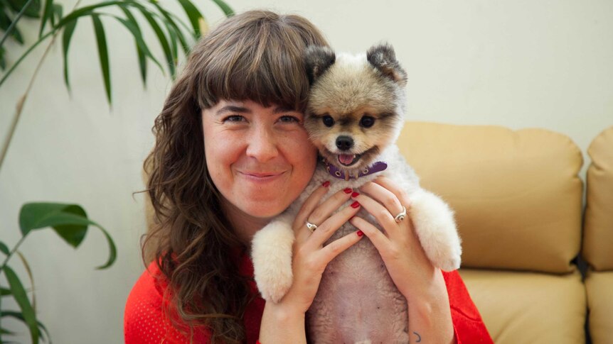 A young woman with a blunt fringe smiles into the camera, holding a chihuahua cross dog close to her face in an embrace.