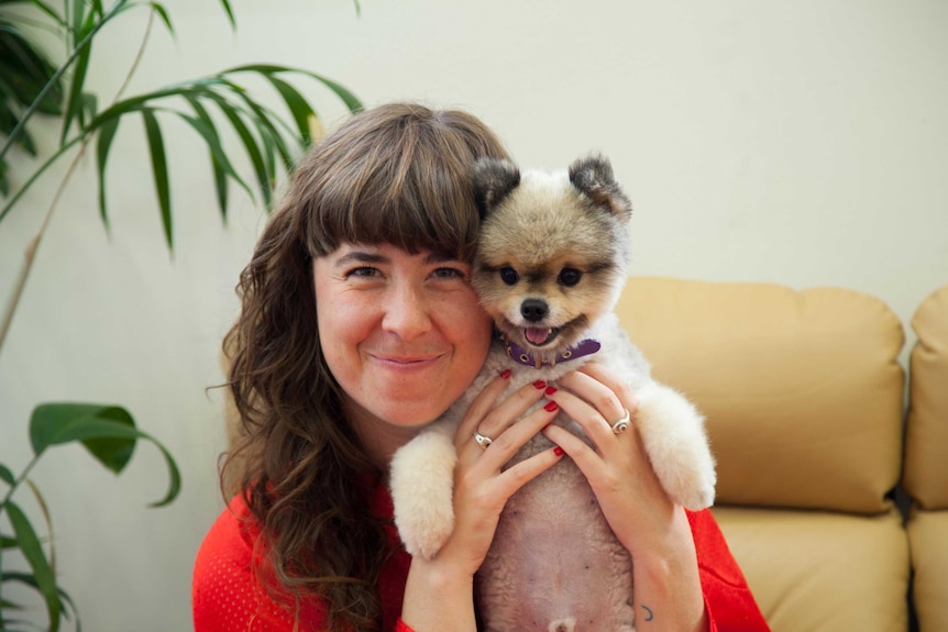 A young woman with a blunt fringe smiles into the camera, holding a chihuahua cross dog close to her face in an embrace.