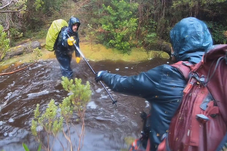 Two men in bushwalking wet weather gear cross a swollen creek in the rain