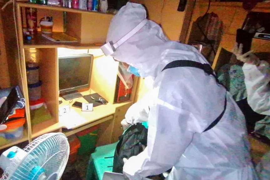 A person in full PPE standing next to a computer in a messy bedroom