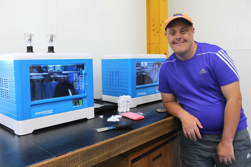 A man leans against a work bench that has two 3D printers operating on it
