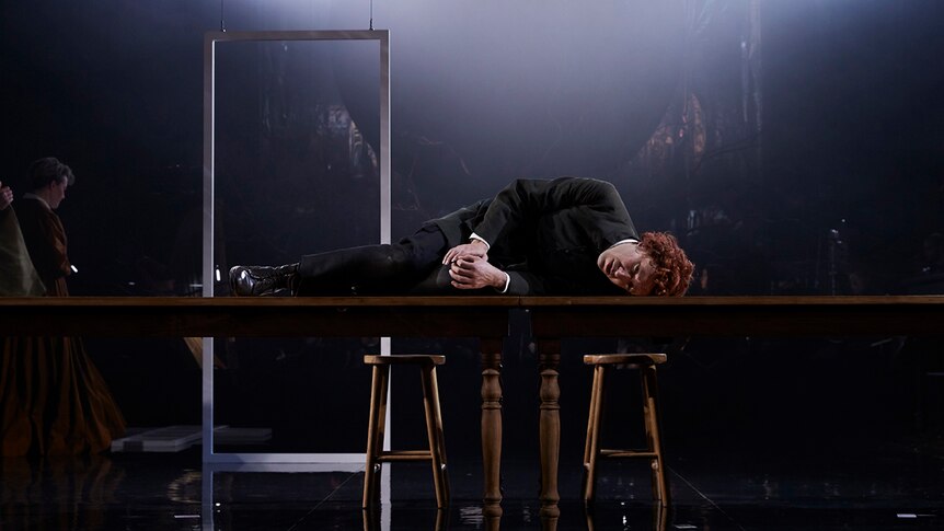 Man with short curly red hair dressed in colonial era costume lying on his side on wooden table on a haze-filled darkly lit set.