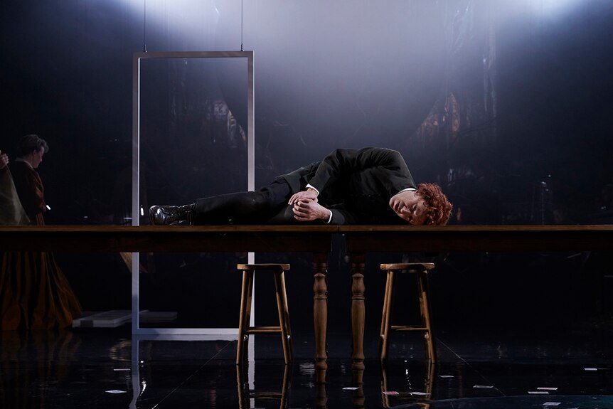 Man with short curly red hair dressed in colonial era costume lying on his side on wooden table on a haze-filled darkly lit set.