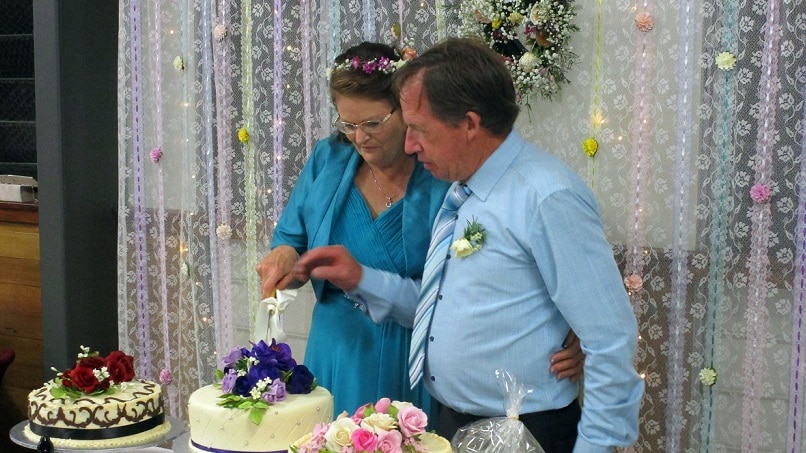 Older couple cuts cake at their wedding, bride is dressed in blue outfit, groom has blue shirt and tie.