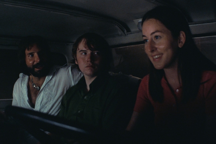 A 40-something man with long hair and a beard, a teenage boy, and a 20-something brunette woman sit together in a truck's cab