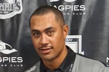 A man in front of a Magpies Sports Club poster smiles at the camera.