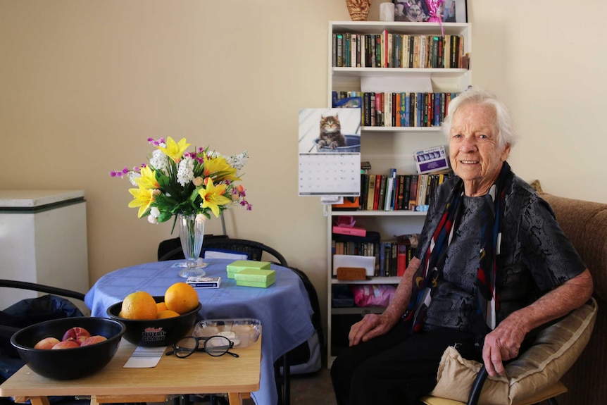86-year-old Laurel Dakin sits in her apartment's dining room with a bookshelf in the background.