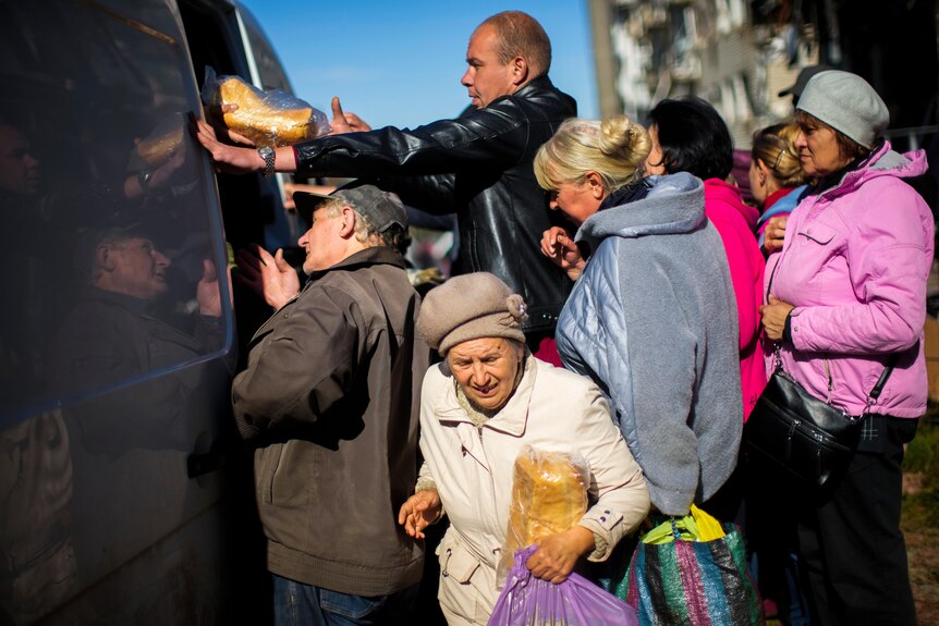 An old lady holds a loaf of bread as a line of people collect food from a vehicle on winter's day