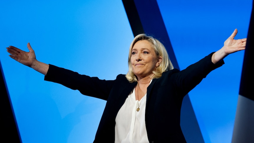 Marine Le Pen stands and spreads her arms out wide while wearing a black jacket wiht a white shirt.