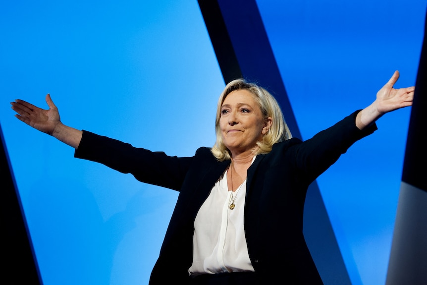 Marine Le Pen stands and spreads her arms out wide while wearing a black jacket wiht a white shirt.