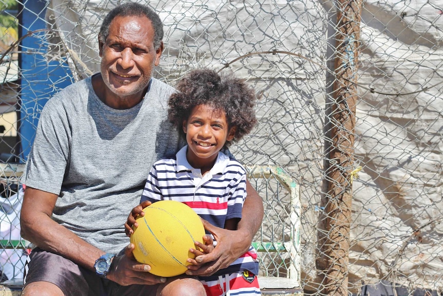 An elderly PNG man sits on a bench with a young boy with wild hair.