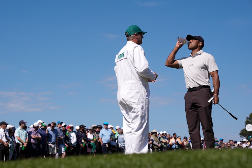 Tiger Woods drinks from a plastic water bottle while standing next to his caddy, in front of a crowd on a golf course.