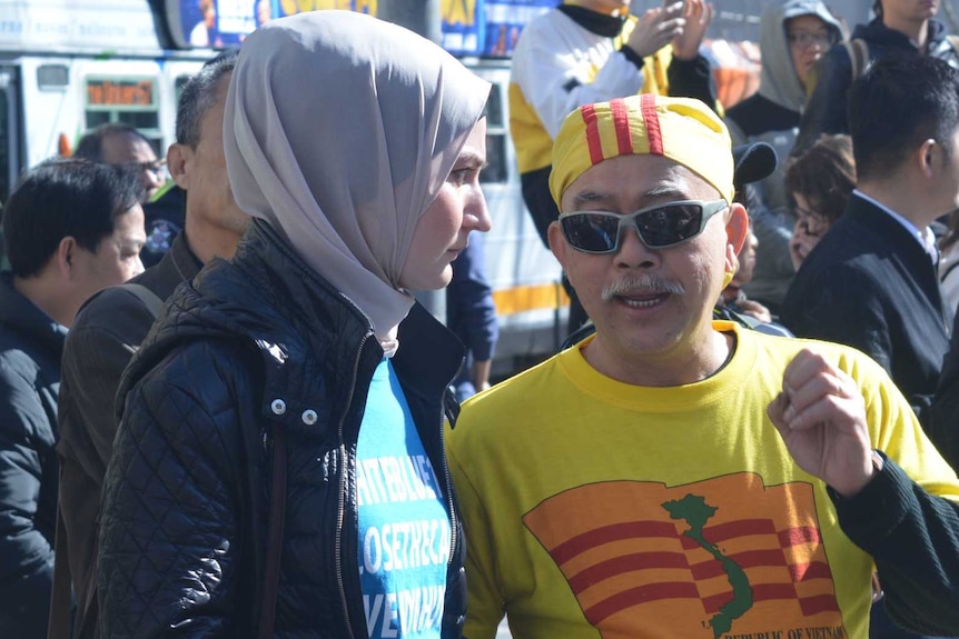 A woman wearing a hijab and black jacked chats to a Vietnamese man among people checking their phones as a tram passes by