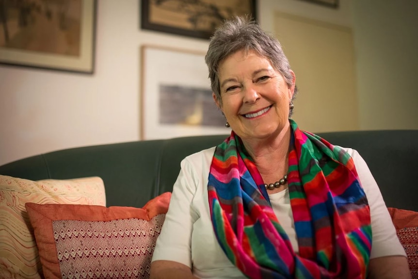 Sue Boyd, with short gray hair, colourful scarf and wide smile, sits on a couch surrounded by several cushions.