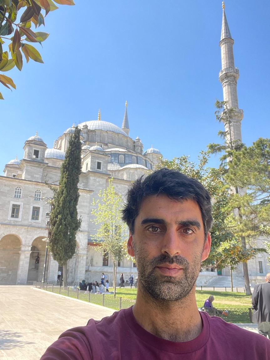 A man with dark hair takes a selfie standing in front of a large Middle Eastern building. 