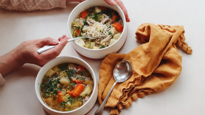 An aerial view of two bowls of vegetable soup with noodles in them, a hand holding a spoon dipping into one, an orange napkin