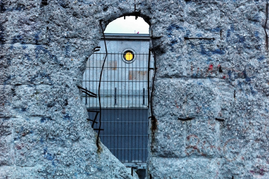 A building can be seen through a hole in a thick concrete wall.