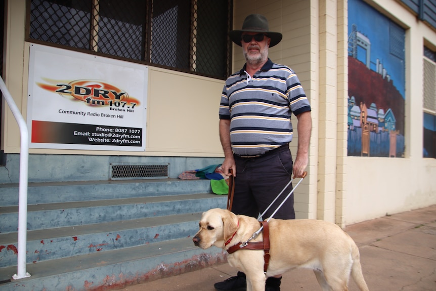 A man wearing a striped shirt, shorts, dark glasses and a hat standing in front of steps and a sign with a guide dog.