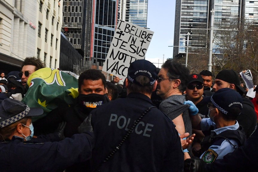 A protest sign reads "I believe in Jesus not the vaccine" as police clash with demonstrators.
