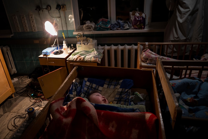 A baby covered in blankets laying in a cot in a rowith a lamp on. 