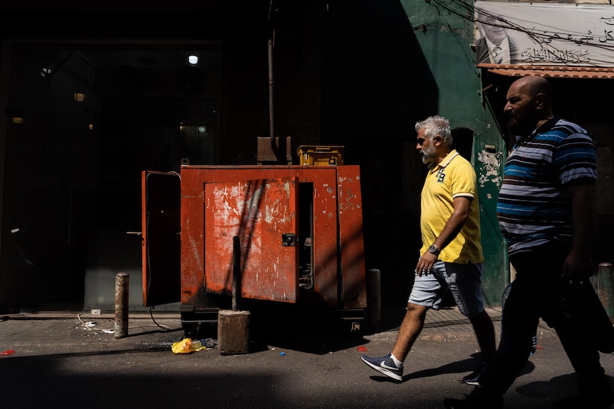 Two men walk past a red electricity generator on a Beirut street 