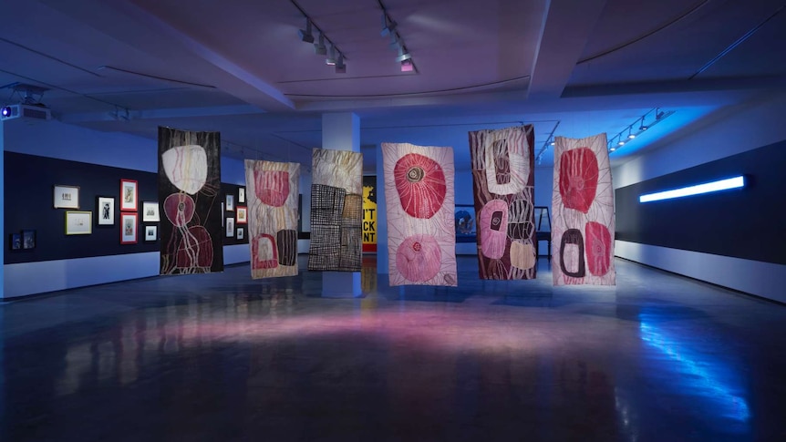 Interior darkened gallery with six bark paintings suspended from the ceiling.