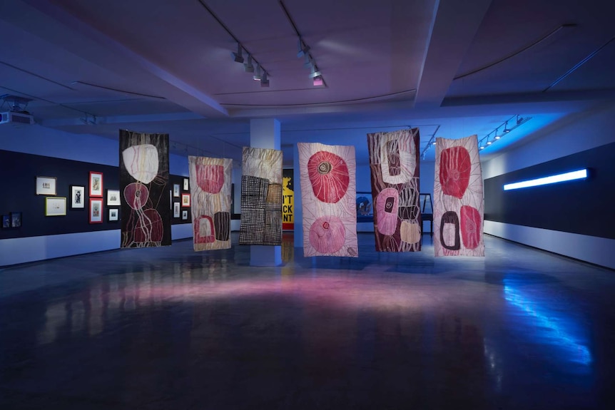 Interior darkened gallery with six bark paintings suspended from the ceiling.