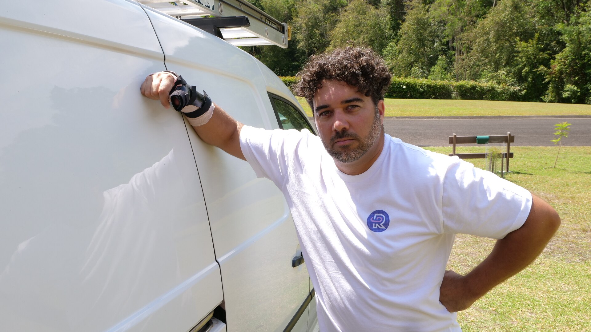 Um homem de cabelos escuros e encaracolados está encostado em uma van estacionada em uma área suburbana.