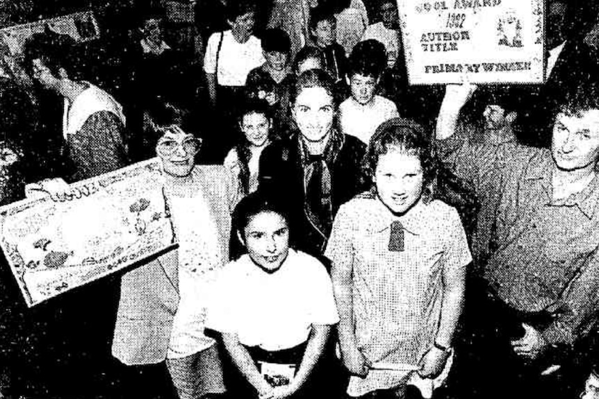 Black and white photo of a man and a woman holding up certificates beside schoolkids