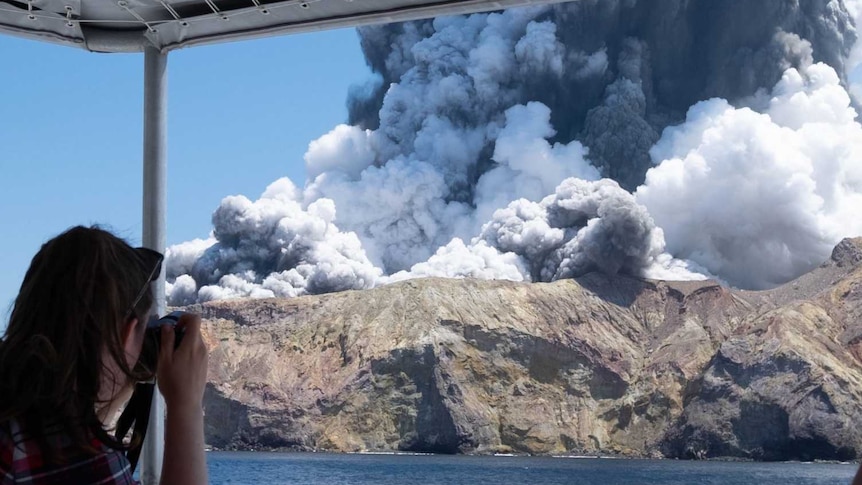 A woman on a boat takes a photo of a volcanic eruption.