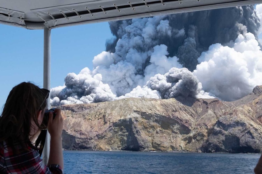 A woman on a boat takes a photo of a volcanic eruption.