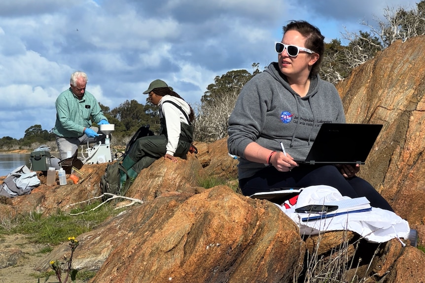 Three researchers sit on rocks by a lake, one has a laptop and a NASA hoodie