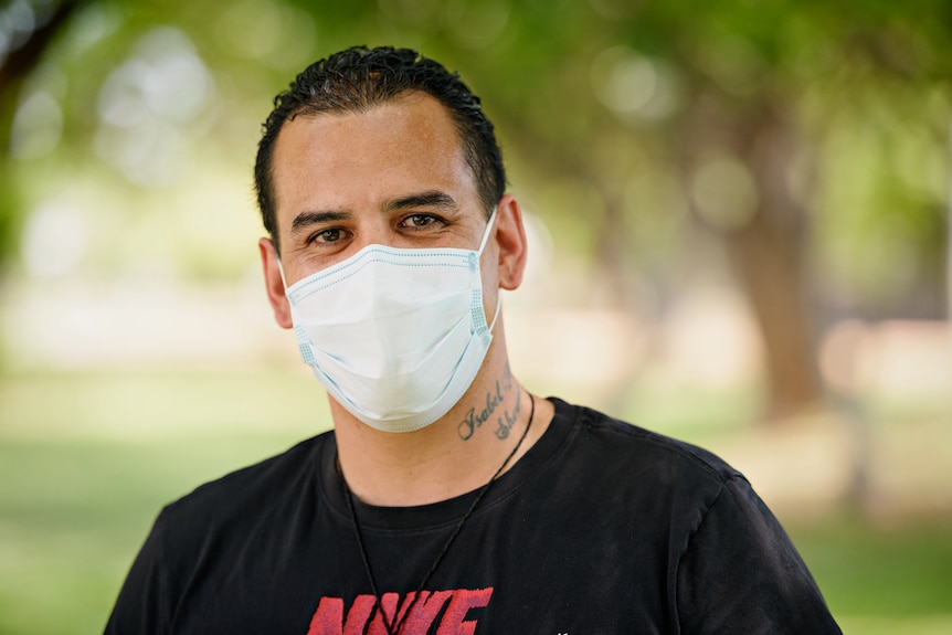 A close-up of a man wearing a surgical face mask, with greenery in the background. 
