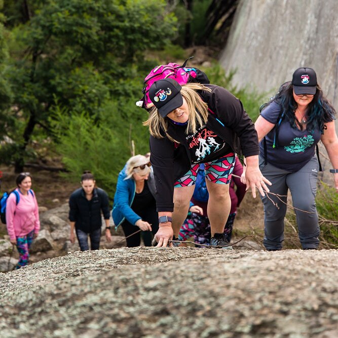 Five women in activewear ascend a large boulder in a scenic forest