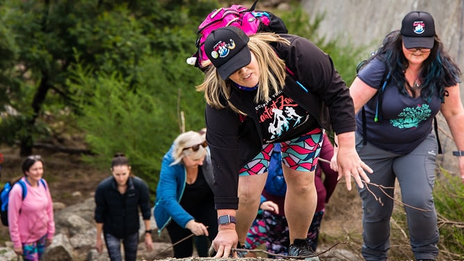 Five women in activewear ascend a large boulder in a scenic forest