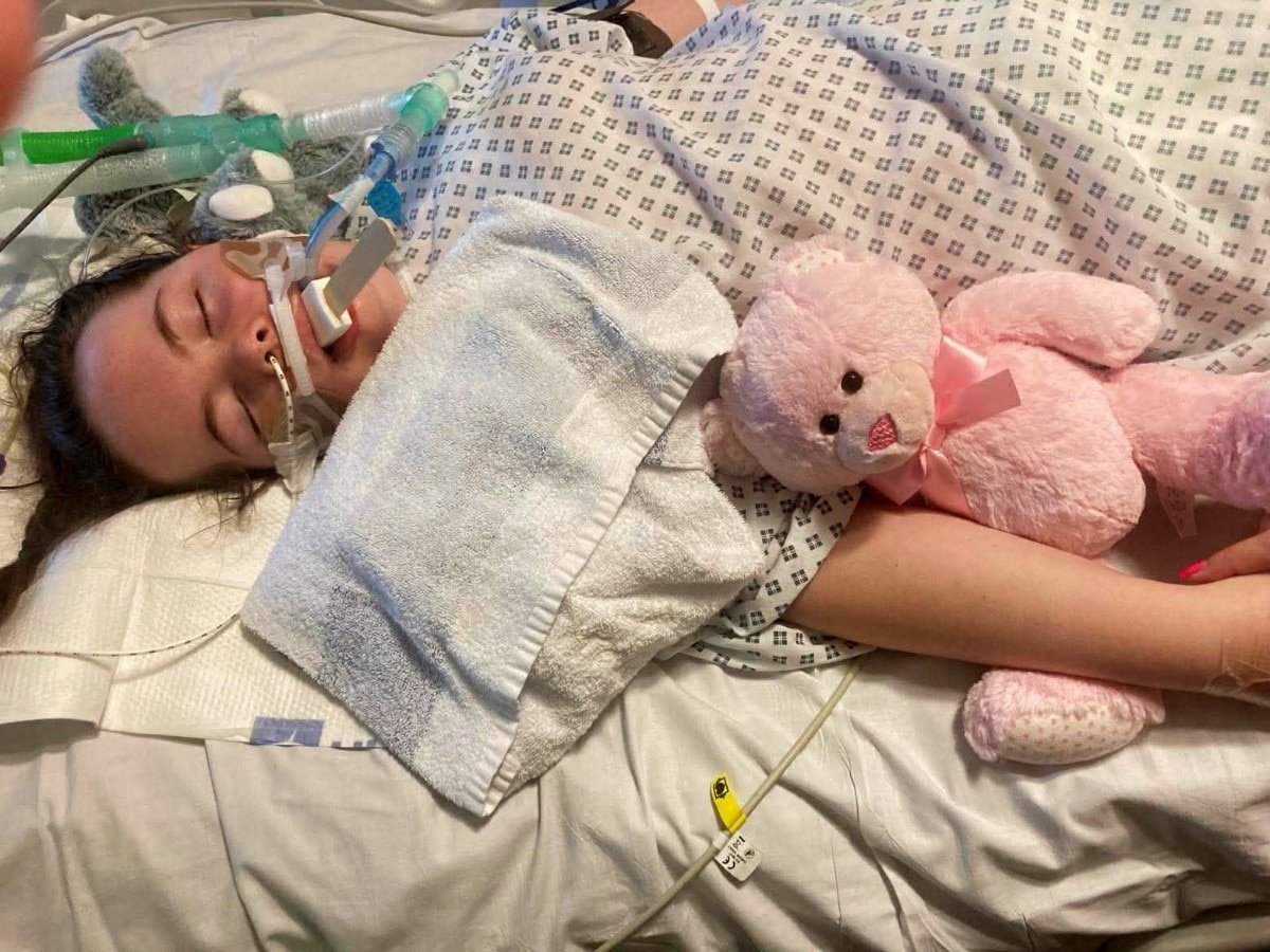 Una niña, con los ojos cerrados y varios dispositivos médicos a su alrededor, en la cama con el brazo alrededor de un osito de peluche. 