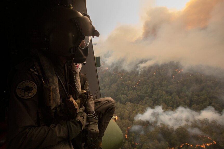 An ADF person looking out of an open helicopter onto smoking bushland.