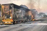 A large burnt out yellow truck is pictured on the side of a road, with a spill on the ground and smoke and flames around