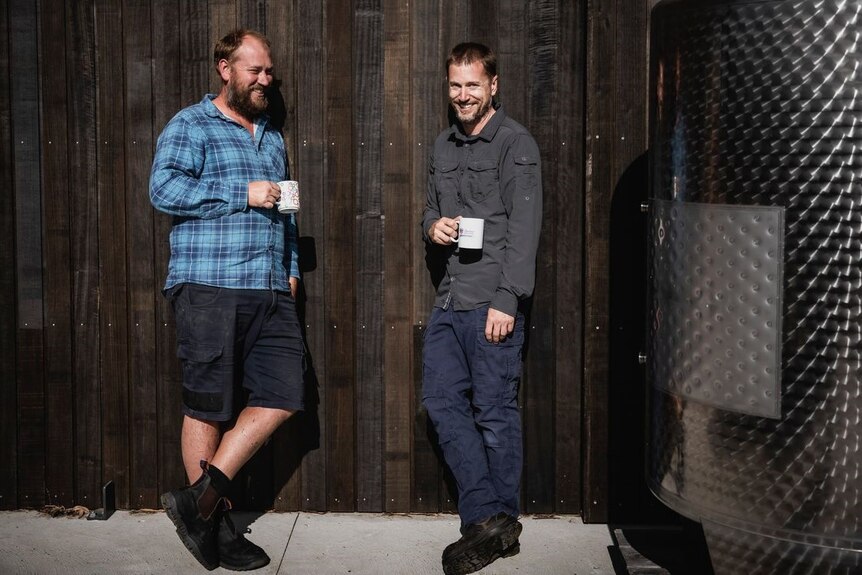 two men stand next to a wine tank holding coffee mugs